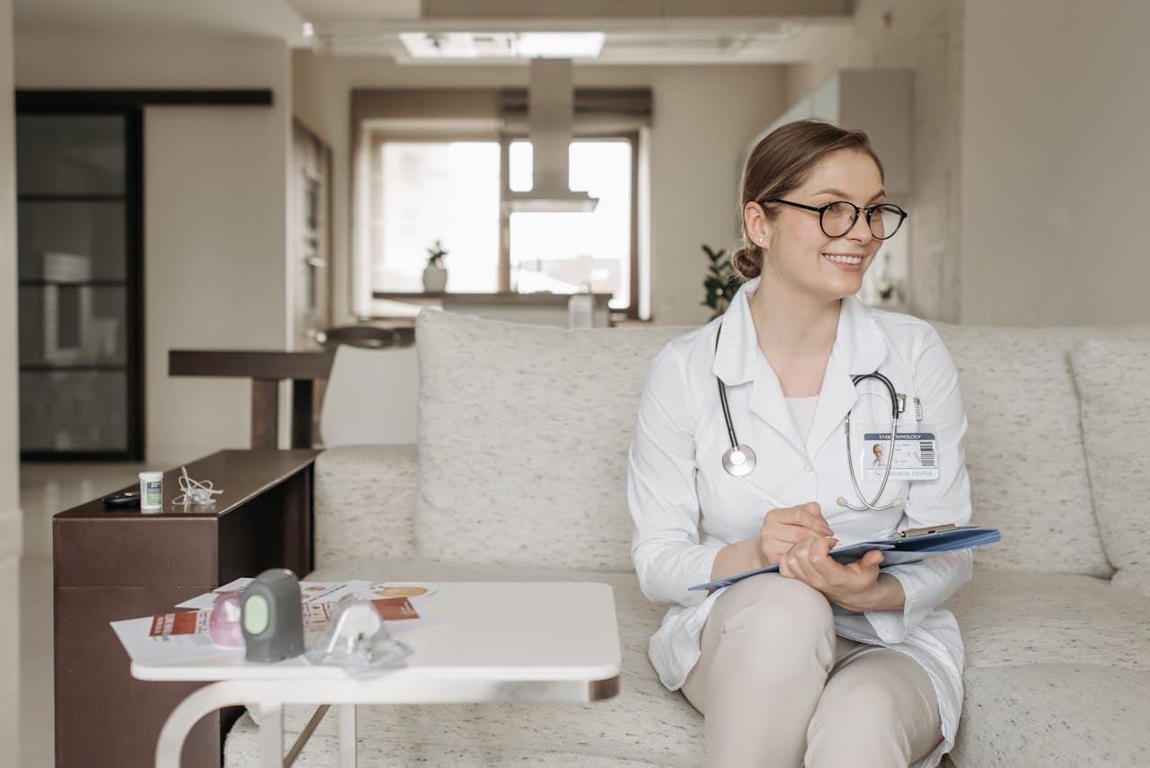 Smiling female doctor on sofa with clipboard and stethoscope in modern office setting.